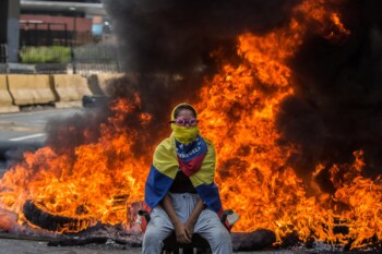 A young hooded woman sitsin front of a barricade fireduring the day of the nationalsit-in called by theVenezuelan opposition. 2017.Caracas, Venezuela